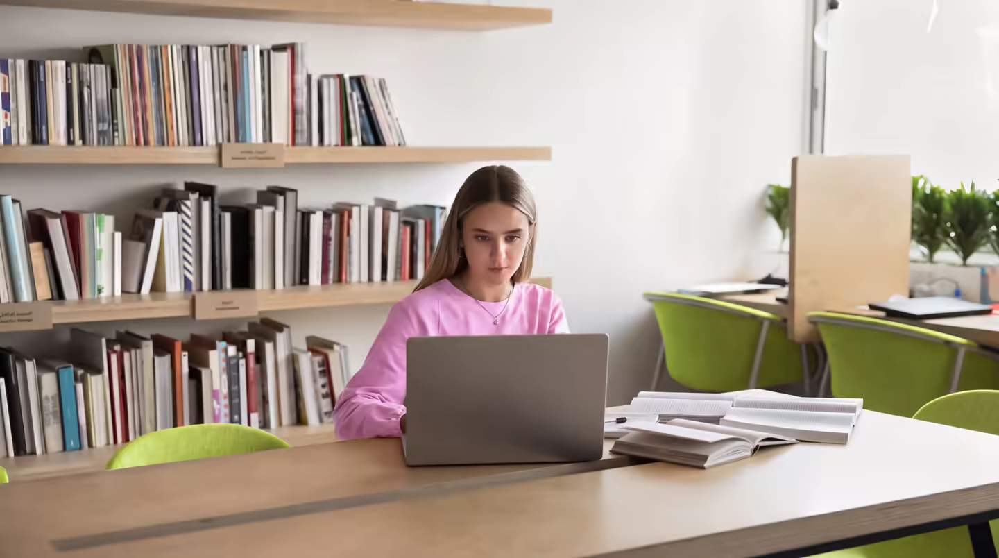 A student working at a laptop at a library desk, books and papers beside her.